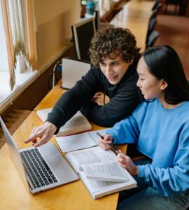 Two students working together on a laptop at a desk in a library, smiling and engaging.